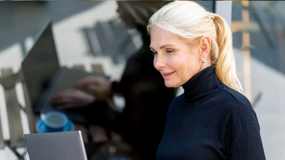 Ältere Frau mit blondem Haar arbeitet lächelnd an einem Laptop im Freien