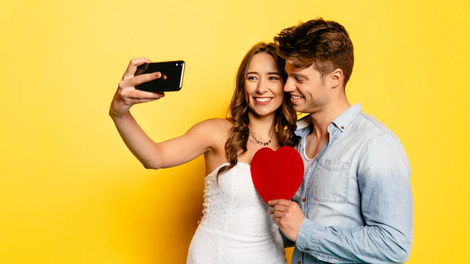 Junges Paar macht ein Selfie mit rotem Herzpapier vor gelbem Hintergrund