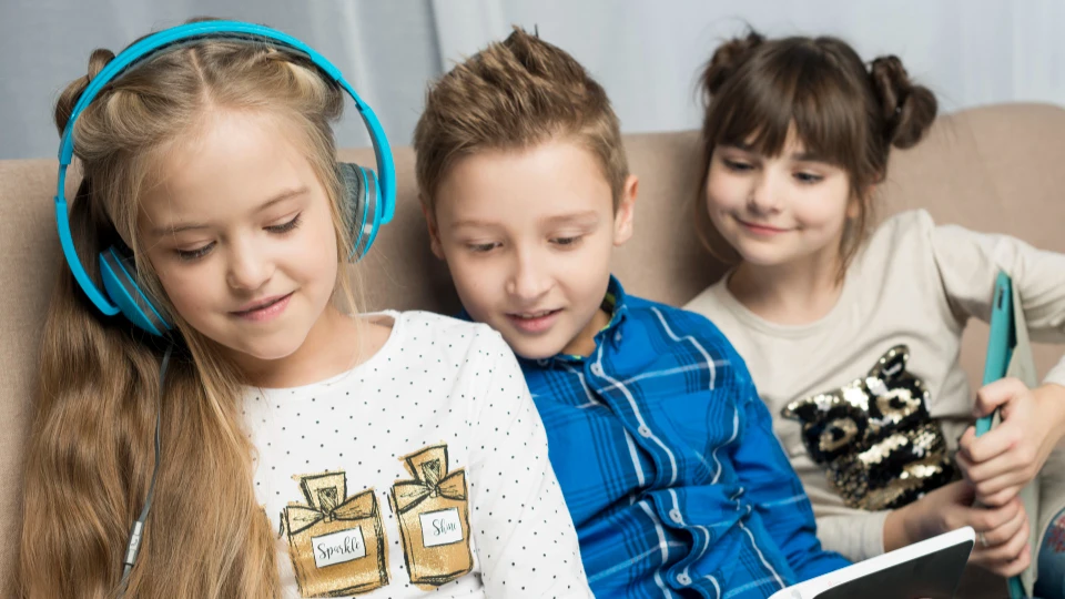 Drei Kinder mit Kopfhörern und Tablet sitzen gemeinsam auf einem Sofa