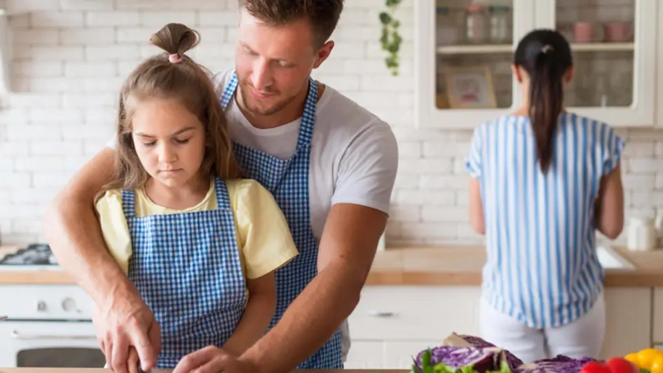 Vater bringt seiner Tochter das Kochen bei während die Mutter im Hintergrund am Herd steht