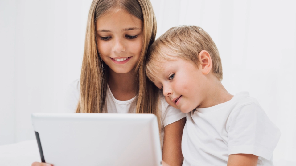 Mädchen und kleiner Junge schauen gemeinsam lächelnd auf ein Tablet vor weißem Hintergrund