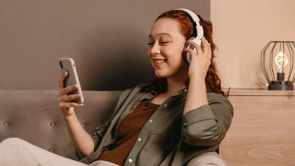 Junge Frau liegt entspannt auf dem Sofa und hört mit Kopfhörern Musik über ihr Smartphone