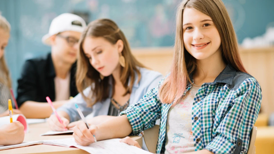 Lächelndes Teenager-Mädchen schaut in die Kamera während Mitschüler im Hintergrund schreiben